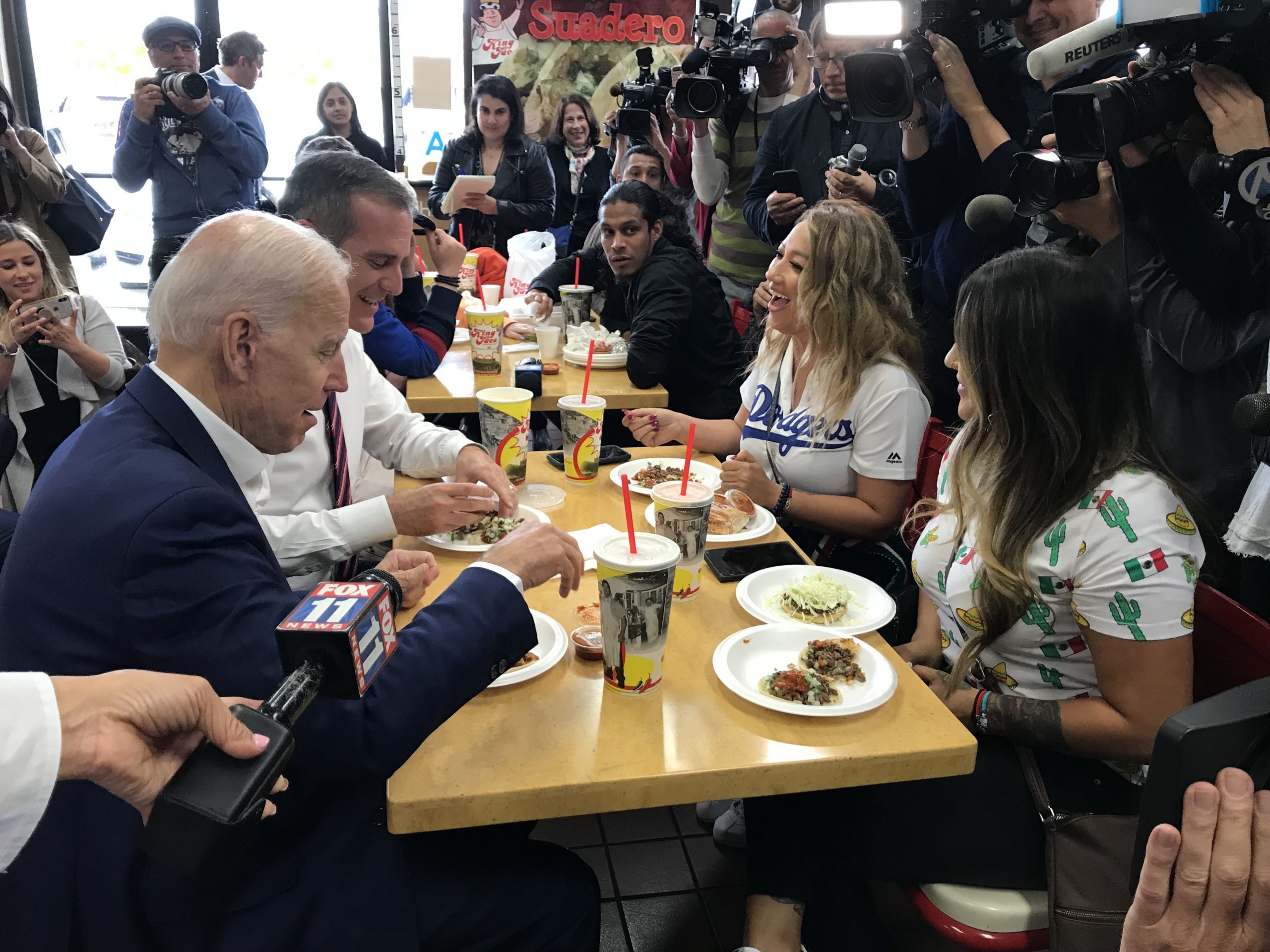 A group of people, including an elderly man in a blue suit, are seated at a fast-food restaurant table, eating and talking. Several Berkeley Journalism personnel with cameras surround them, capturing the moment. Food trays with drinks and various dishes are on the table.