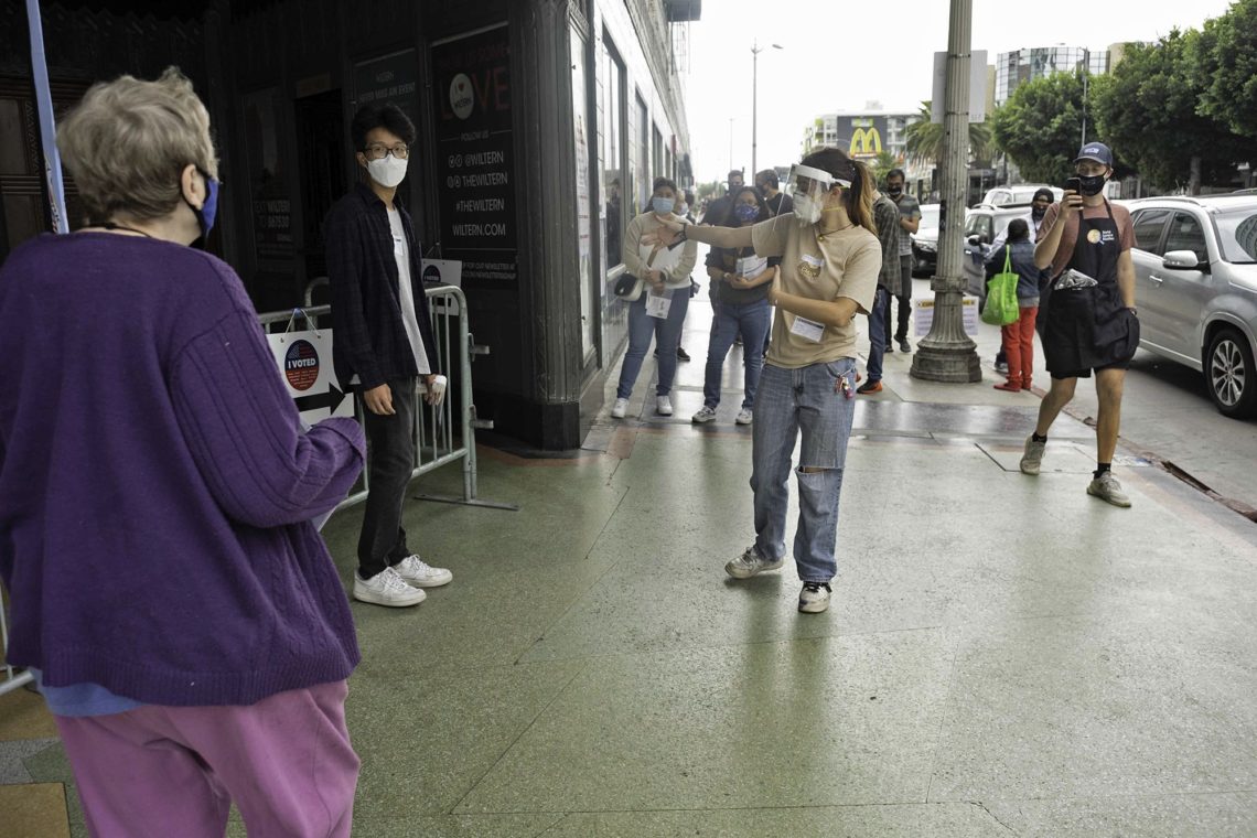 A group of people wearing masks stand and maintain social distance in line on a sidewalk. An individual in a cap and mask appears to be directing the crowd. The background includes storefronts, a McDonald