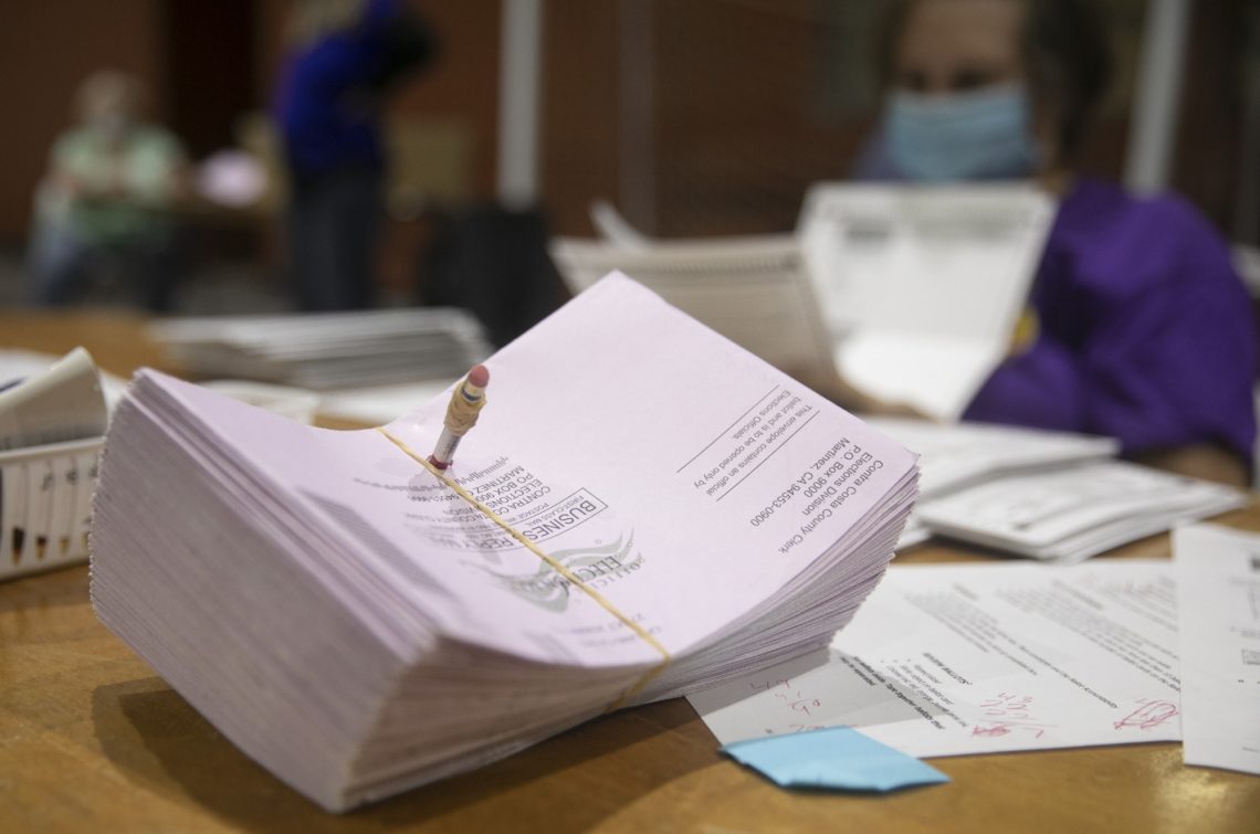 A stack of pinkish ballots held together with a rubber band lies on a table. In the background, a person wearing a face mask and purple shirt appears to be sorting through additional papers. The setting suggests a vote counting or ballot processing environment, reminiscent of Berkeley Journalism
