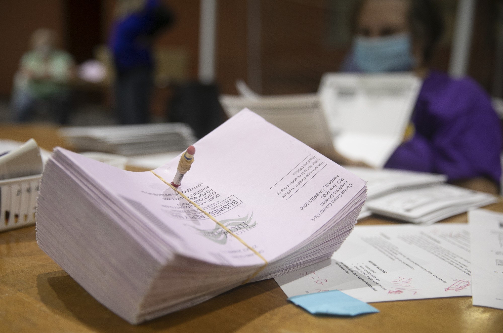 A stack of pinkish ballots held together with a rubber band lies on a table. In the background, a person wearing a face mask and purple shirt appears to be sorting through additional papers. The setting suggests a vote counting or ballot processing environment, reminiscent of Berkeley Journalism's detailed election coverage.
