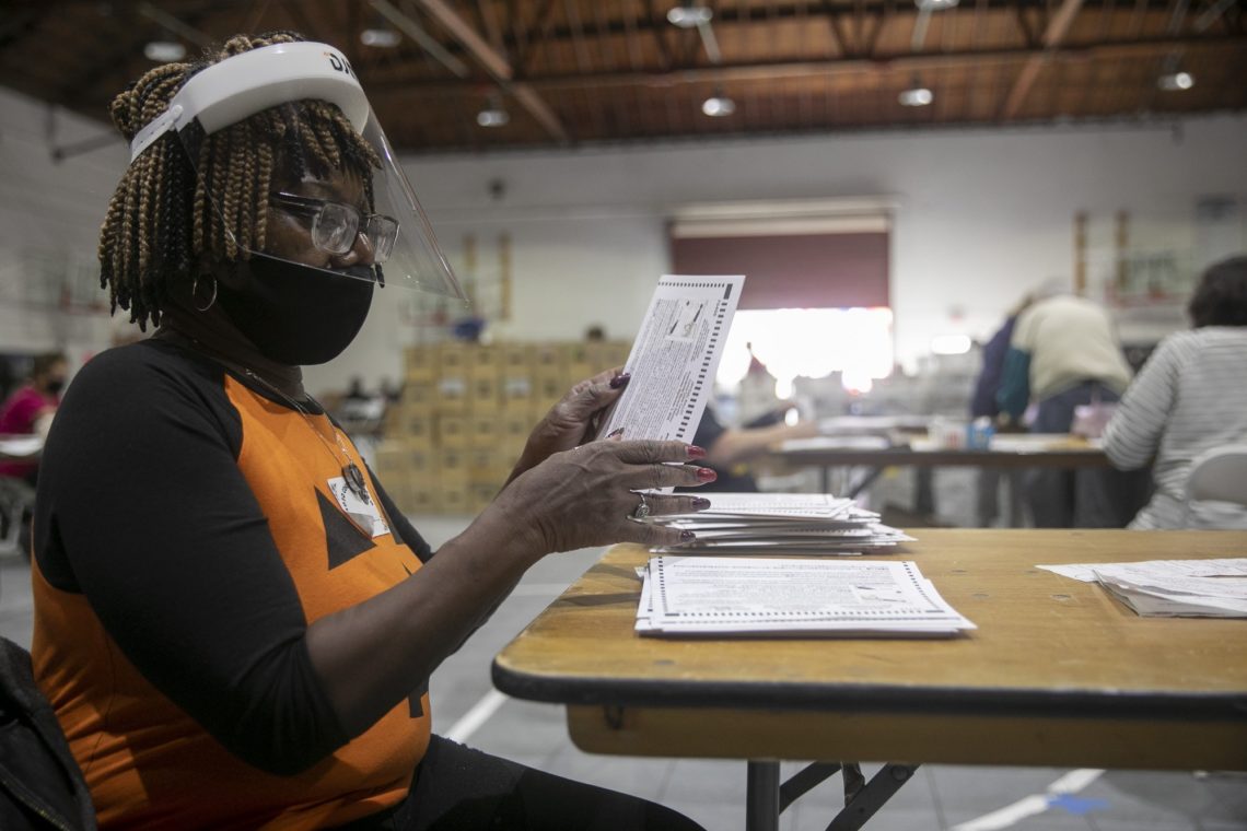 A woman wearing a face shield, mask, and orange shirt is seated at a table sorting through stacks of paper ballots in a large room. It