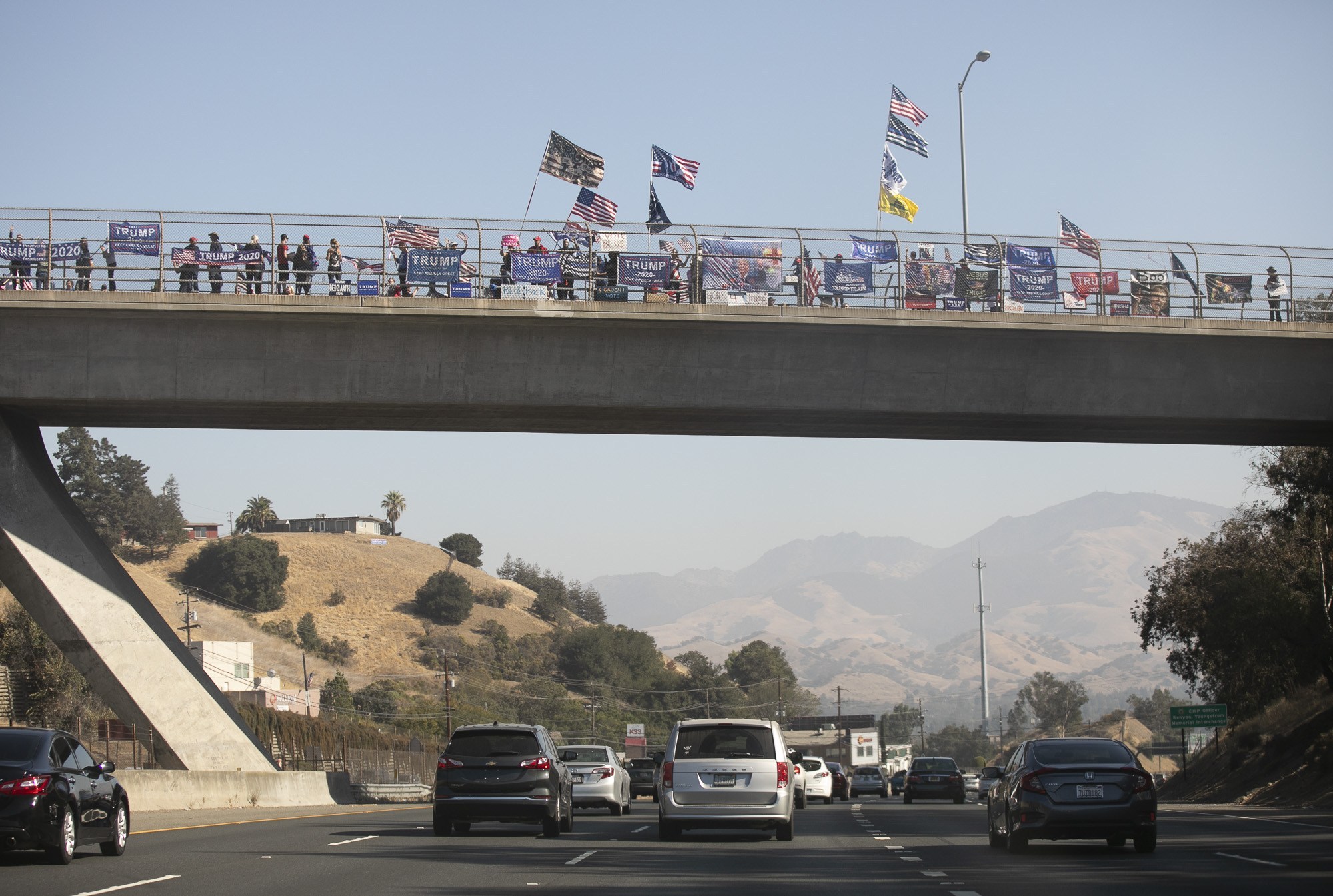 A group of people stand on a highway overpass holding flags and signs in support of a political rally. Several American flags and banners are displayed. Cars are visible on the highway below, and mountains can be seen in the background under a clear sky. A Berkeley Journalism project captures this moment.