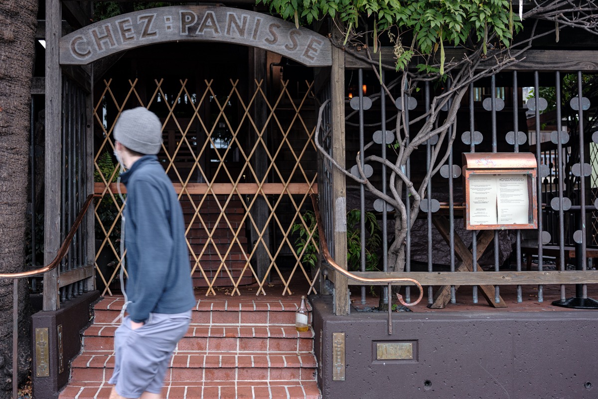 A person in a grey beanie and dark jacket walks past the closed entrance of Chez Panisse, a restaurant with a wooden exterior and stairs leading up to a doorway blocked by a wooden gate. A menu is displayed on the right side near the door, capturing what could be Berkeley Journalism at its finest.