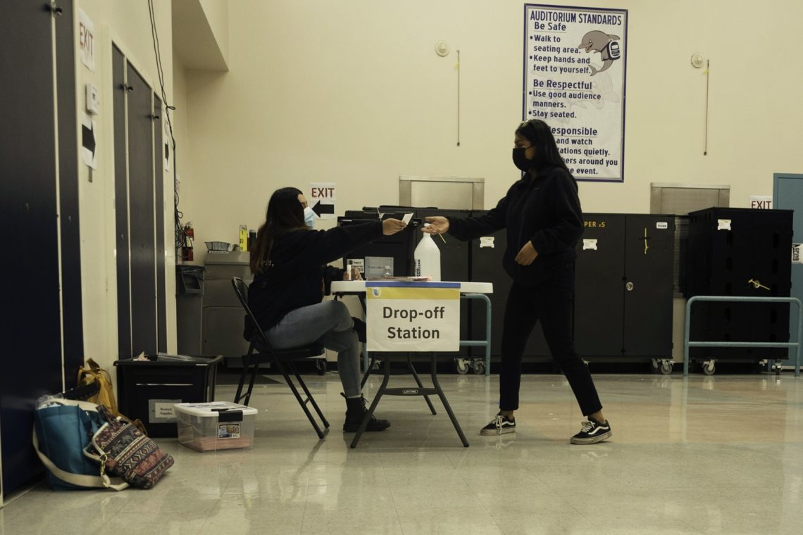 Two individuals interact at a "Drop-off Station" in a spacious indoor setting. One person, seated at a table with the sign “Drop-off Station,” hands an item to another person standing nearby. Both wear masks. Signs with guidelines hang on the wall in the background, reflecting Berkeley Journalism