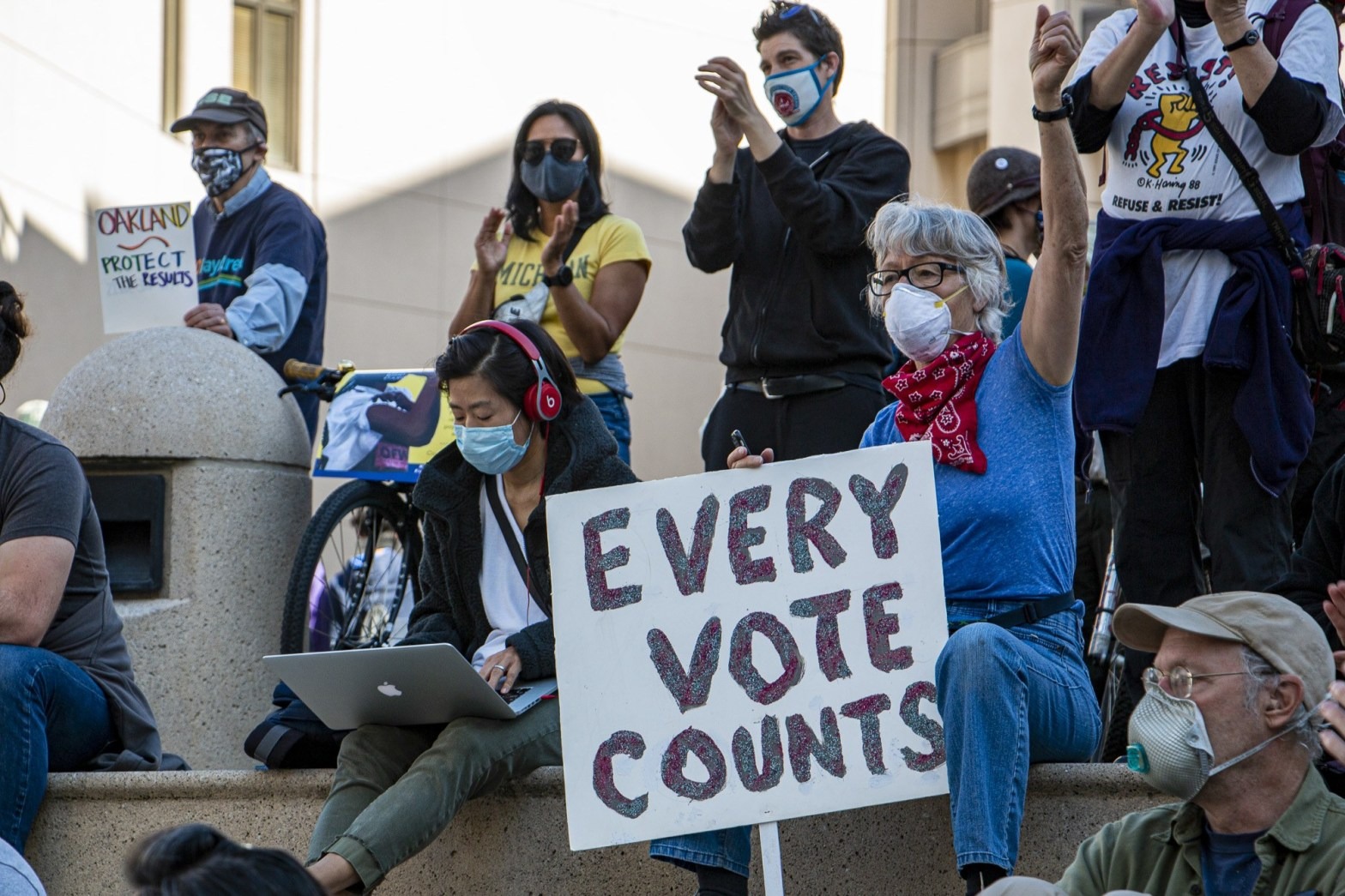 A group of diverse people, some wearing masks, gather outdoors. A seated person holds a sign that reads, 