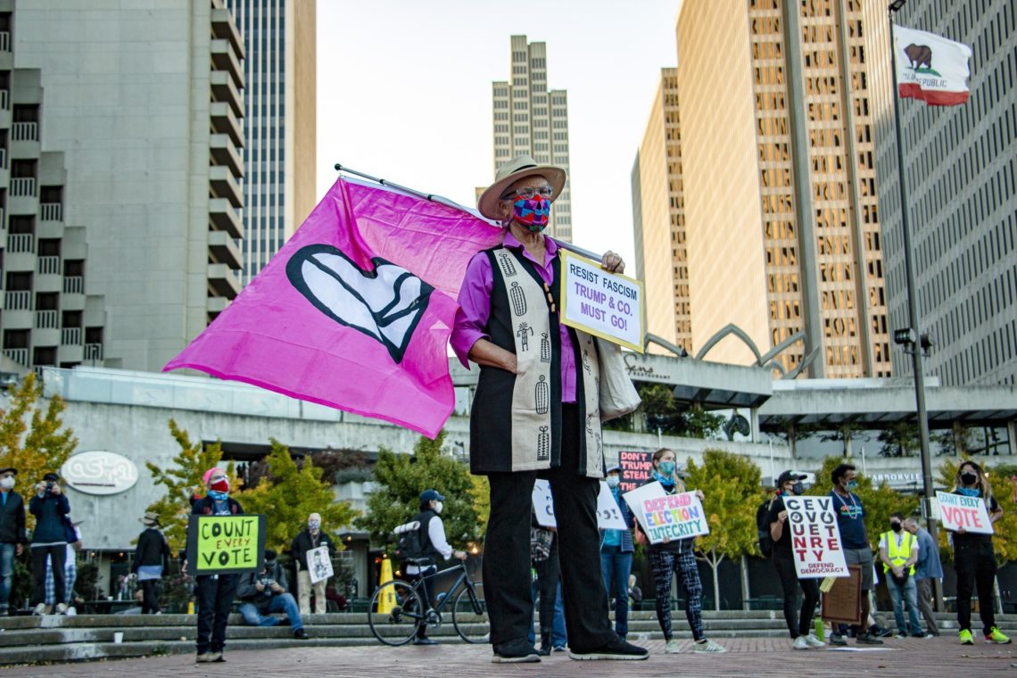 Person holding a large pink flag with a heart symbol, standing in an urban area with tall buildings. Surrounded by others holding protest signs, including "Count Every Vote" and "Trump & Co Must Go." Many wear face masks, and trees are visible in the background—captured perfectly by Berkeley Journalism students.