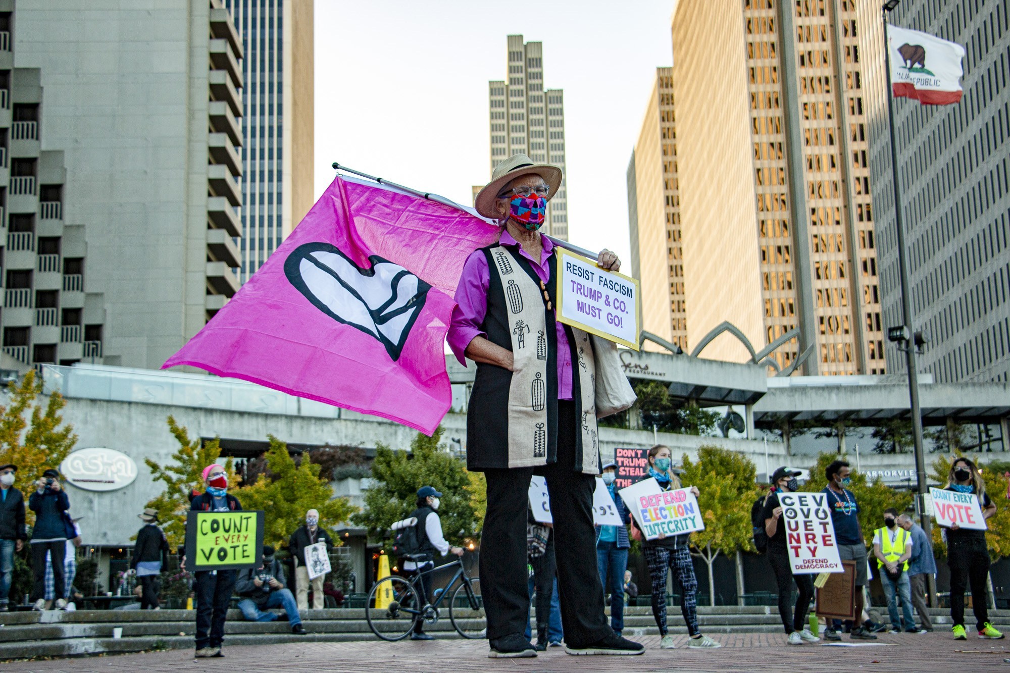 Person holding a large pink flag with a heart symbol, standing in an urban area with tall buildings. Surrounded by others holding protest signs, including 