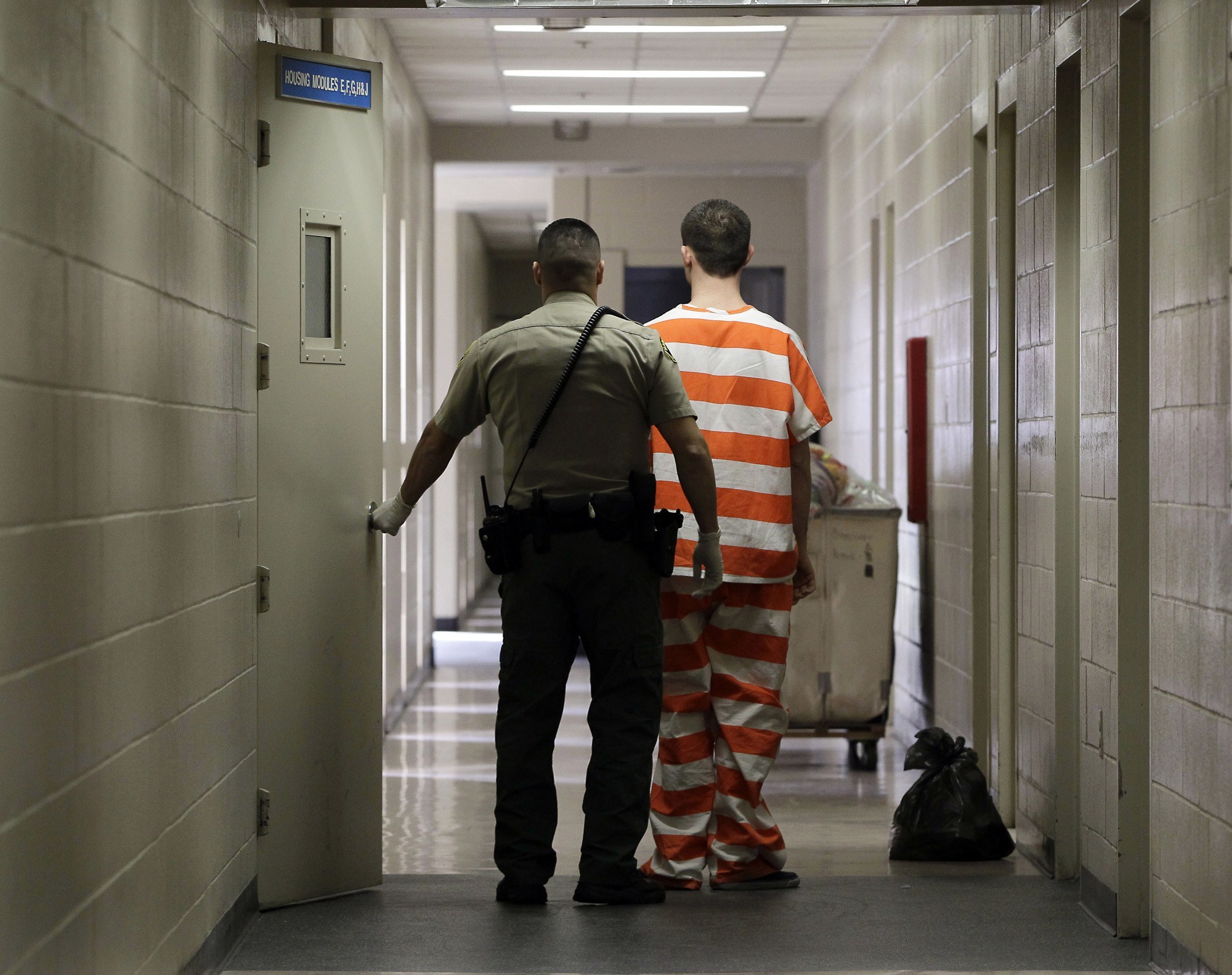 A prison guard holds a door open for an inmate in an orange and white striped jumpsuit. They walk down a long, dimly lit hallway with light beige walls and tiled floors. The inmate, pushing a cart with a bag beside it, could almost be mistaken for someone whisked away from the hustle of Berkeley Journalism hallways.