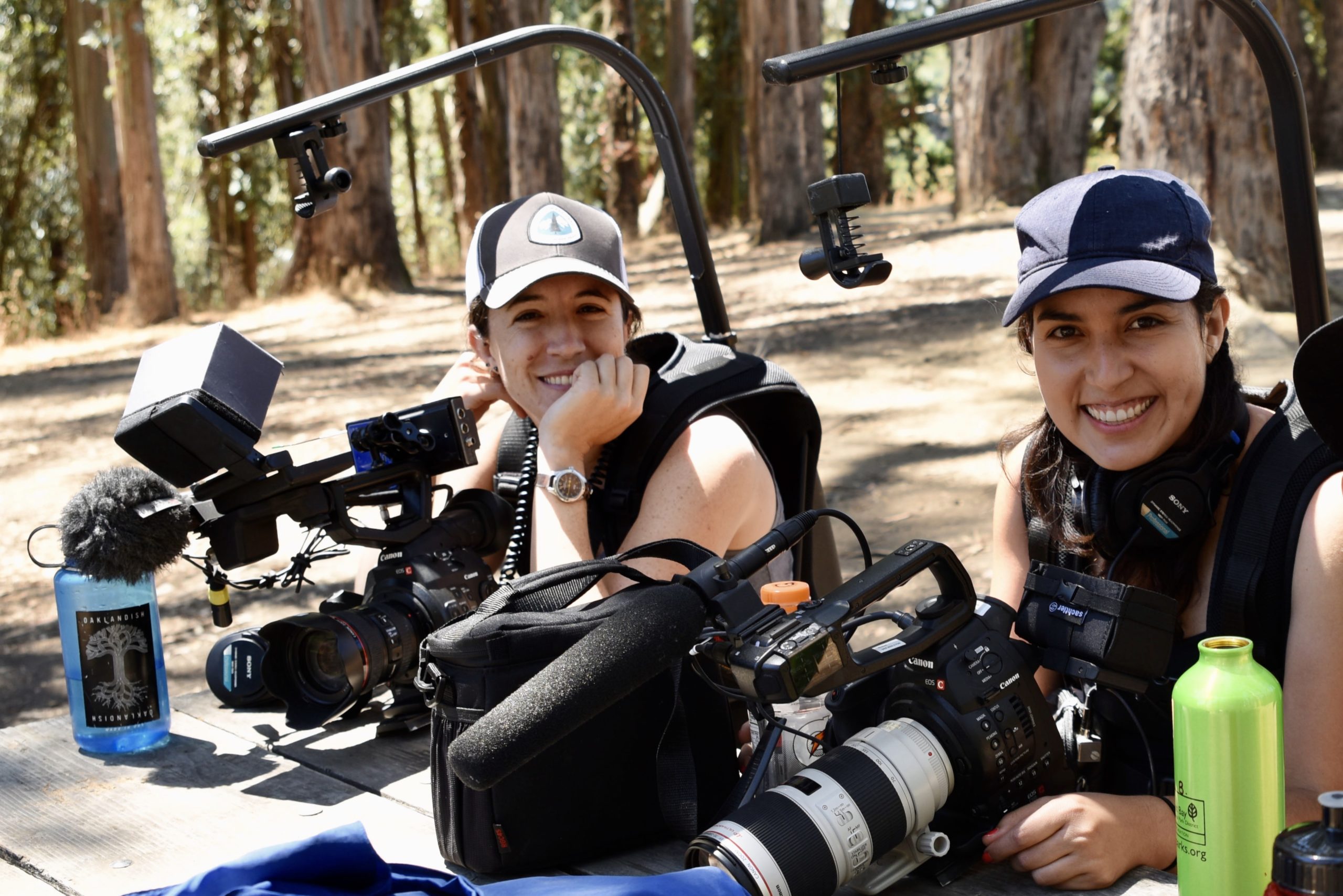 Two smiling individuals wearing hats are seated outdoors with professional camera equipment. Various photography gear, water bottles, and trees in the background can be seen, indicating an outdoor shoot or nature documentation setting. One person holds a camera with a large lens, embodying the Berkeley Journalism spirit.