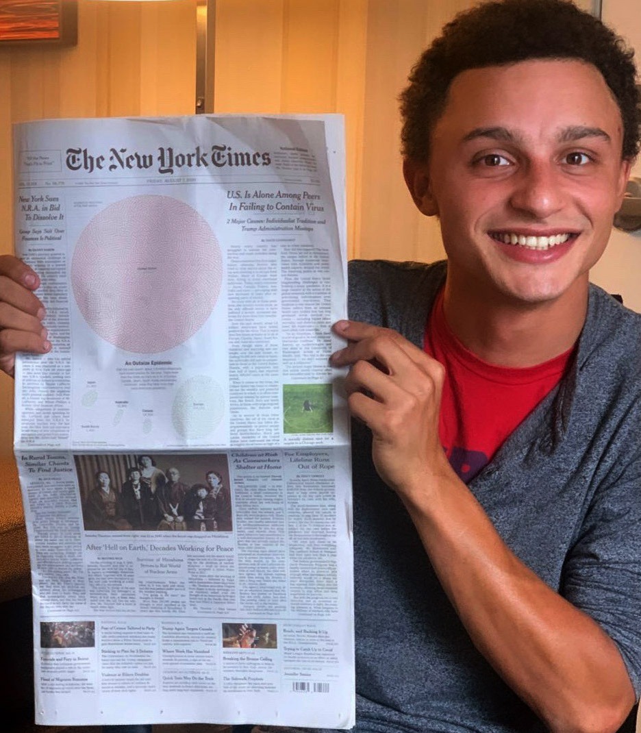 A person with short curly hair, wearing a red shirt and gray cardigan, smiles while holding up a copy of The New York Times newspaper. The front page features a large red circle graphic and an article about the US handling of the coronavirus pandemic, showcasing Berkeley Journalism's insightful reporting.