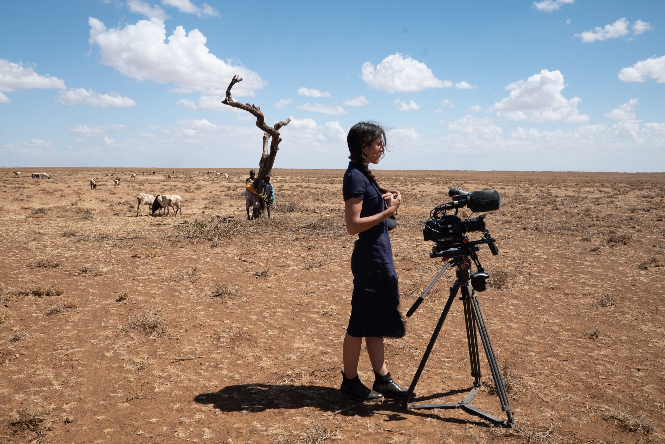 A woman stands beside a video camera on a tripod in an arid, open landscape with a few sparse clouds in the sky. In the background, a leafless tree stands and a person tends to a small herd of goats. The dry, barren terrain serves as an evocative backdrop for this Berkeley Journalism project.