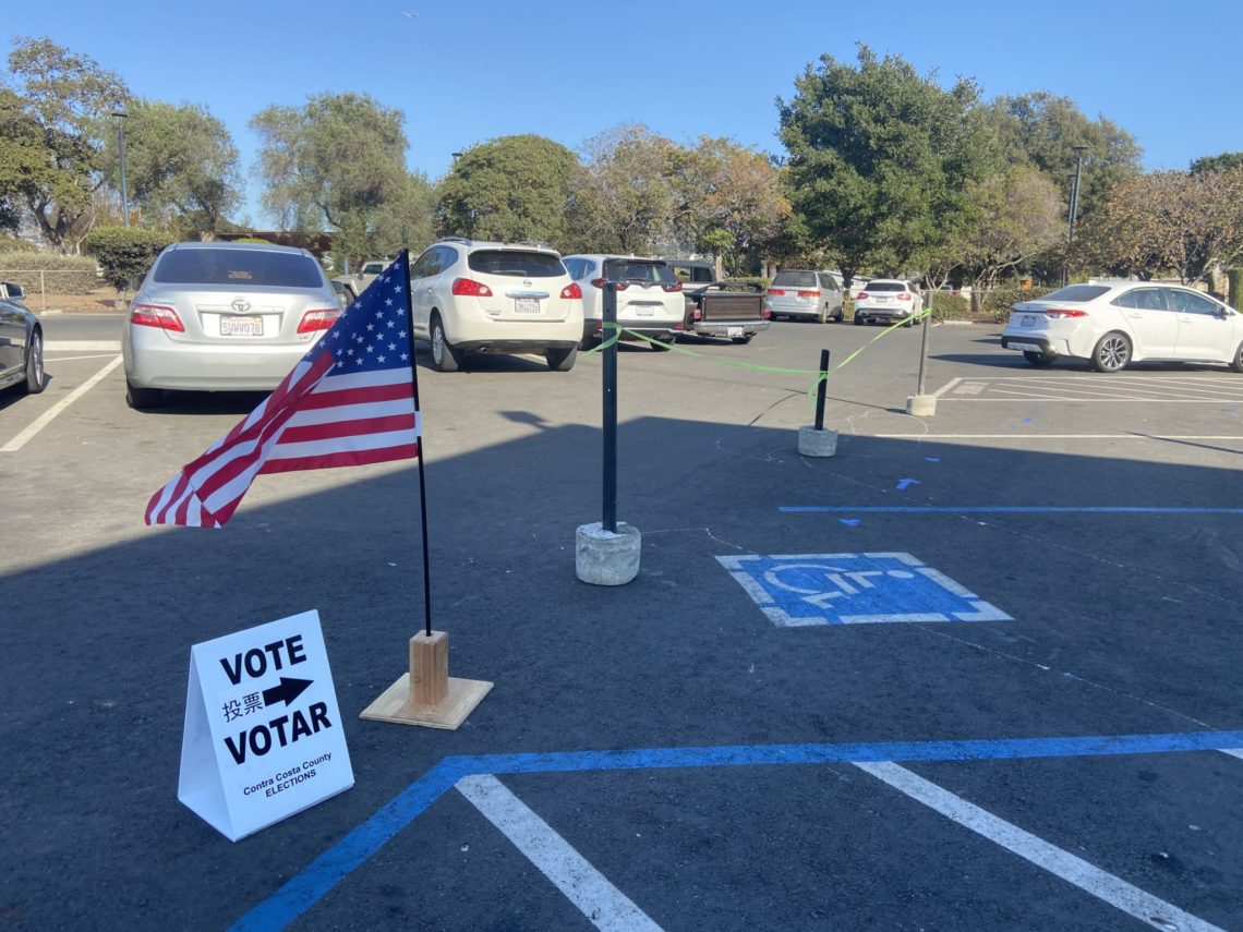 A parking lot with several parked cars on a sunny day. An American flag is planted next to a "VOTE" sign in English, Chinese, and Spanish. The sign includes "Vote Early" and "Vote Center" details. Handicap parking spaces are visible in the foreground—an ideal scene for a Berkeley Journalism feature.
