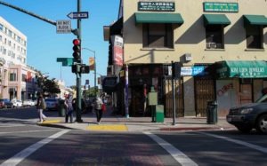 A bustling street corner in Berkeley with various shops, including a jewelry store and a professional services office. Pedestrians cross beneath traffic lights, signs visible. The clear weather bathes the scene in sunlight, casting shadows—an everyday tableau captured by keen Berkeley journalism.