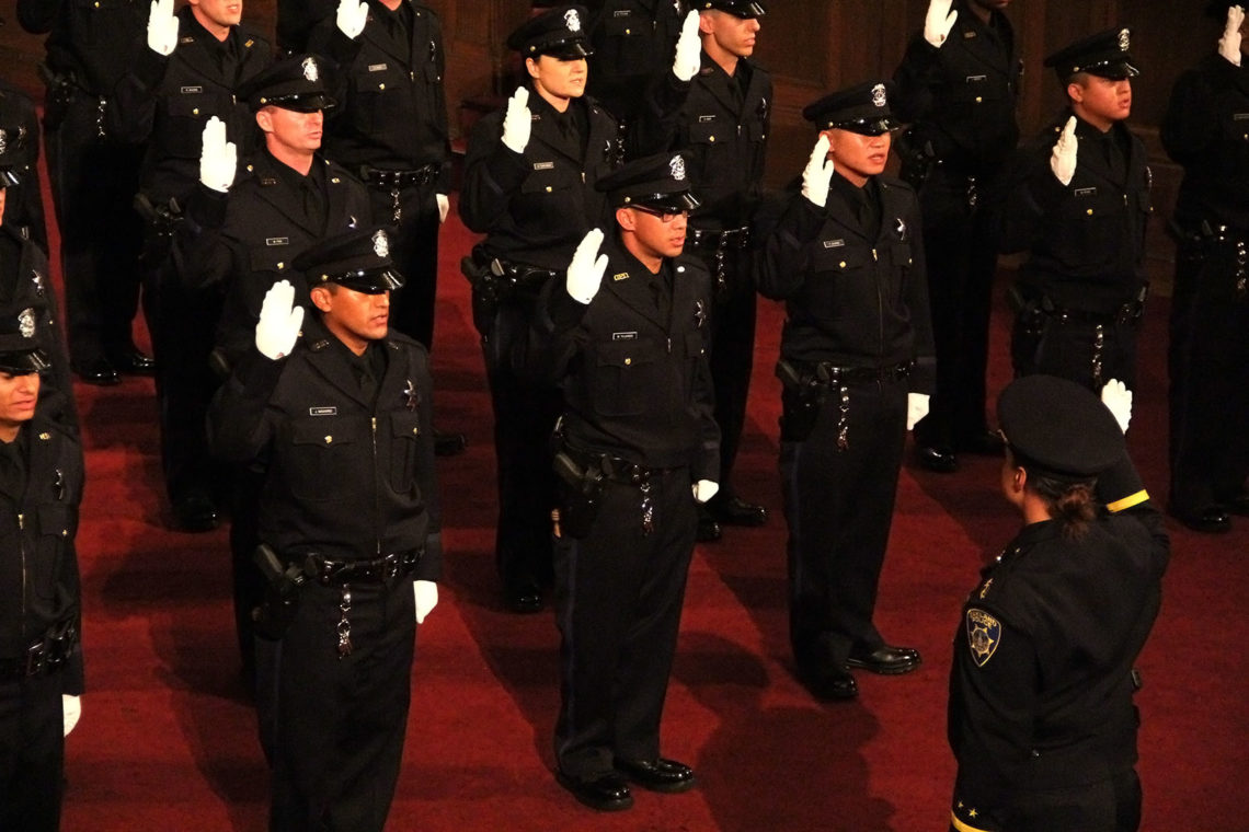 A group of police officers in black uniforms stand with their right hands raised, possibly during a swearing-in ceremony. They are indoors, standing on a red carpet, and another officer in the foreground appears to be leading or supervising the ceremony. Nearby, Berkeley Journalism students capture every moment.