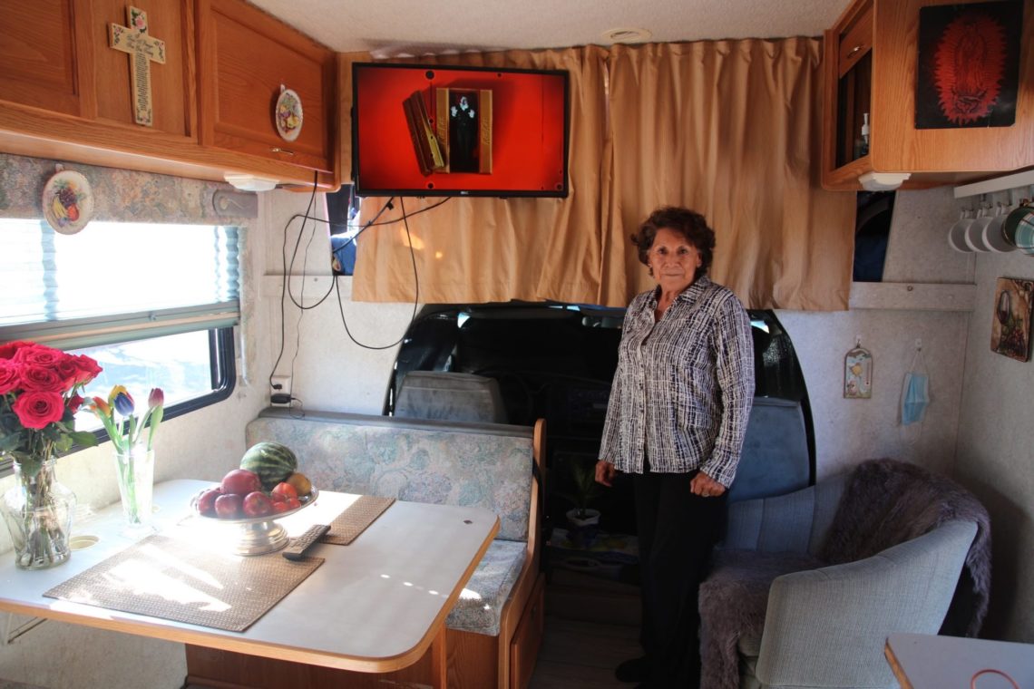 An elderly woman stands inside a cozy, well-lit RV. She is next to a small dining table with a bouquet of red roses and a bowl of fruit. Behind her, the TV mounted on the wall displays Berkeley Journalism news, adding to the charm of the neat and inviting interior decorated with various items.