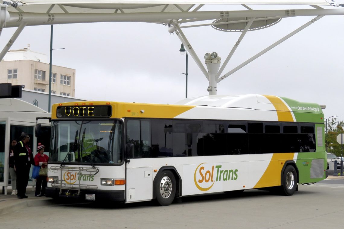 A SolTrans bus is parked at a station with passengers boarding. The bus is white with green and yellow accents, branded with the SolTrans logo. Its destination sign reads "VOTE." In the background, a covered bus stop with supporting structures evokes a scene perfect for a Berkeley Journalism feature.