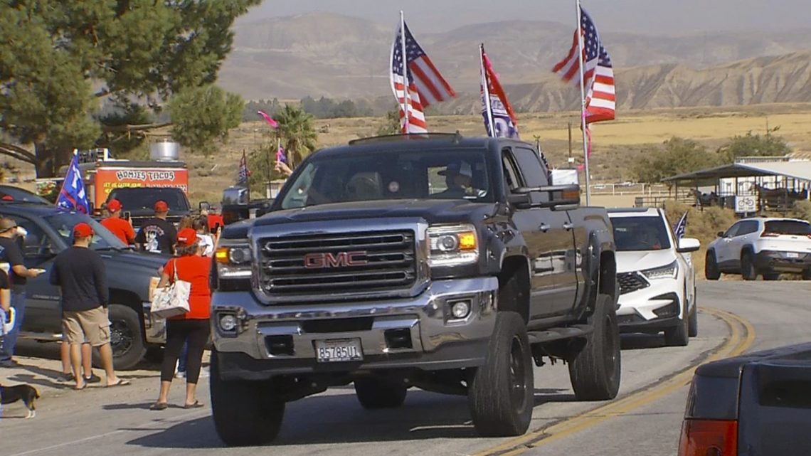 A lifted black GMC truck with several American flags drives down a road as a crowd of people nearby watches. The scene, reminiscent of stories covered by Berkeley Journalism, unfolds in a rural area with mountains in the background. Other vehicles and a food truck are also visible.