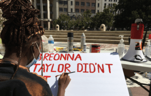 A person with braided hair and a mask paints "BREONNA TAYLOR DIDN'T GET" in red letters on a white poster. They are seated at an outdoor table with various art supplies, spray paint, and a megaphone nearby. Buildings and trees are visible in the background, epitomizing the spirit of Berkeley Journalism.