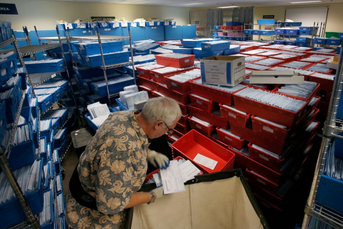 A worker in a patterned shirt sorts through a large number of mail-in ballots inside a room filled with blue and red plastic bins, each containing stacks of envelopes and papers. The room, reminiscent of the organized chaos often reported by the meticulous eyes at Berkeley Journalism, has shelves on the sides filled with mail.