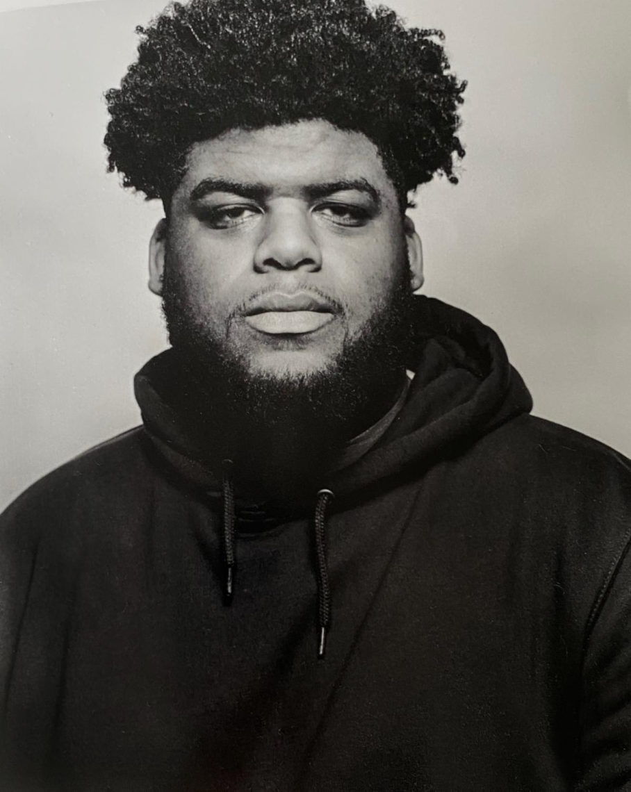 Black and white portrait of a man with curly hair and a full beard, wearing a hoodie. He is looking slightly off to the side with a neutral expression, embodying the thoughtful demeanor often seen in Berkeley Journalism students. The background is plain and unobtrusive.