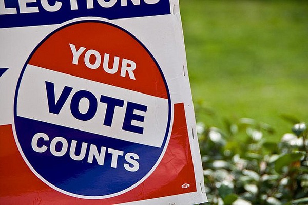 A colorful election sign reads "Your Vote Counts" in bold letters, with a red, white, and blue color scheme. The sign stands outdoors amidst green grass and foliage. Capturing the essence of civic duty, it