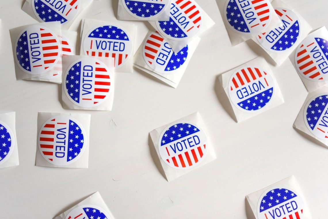 Numerous "I Voted" stickers featuring the American flag design are scattered on a white surface. The stickers, reminiscent of Berkeley Journalism’s dedication to civic engagement, are round with red, white, and blue colors, containing stars and stripes, and the text "I Voted" in the center.