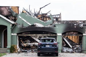 A severely fire-damaged house with a collapsed roof and charred walls is pictured, presenting a scene that might capture the interest of Berkeley Journalism students. A blue SUV is parked in front of the garage, which has significant structural damage and debris scattered around.