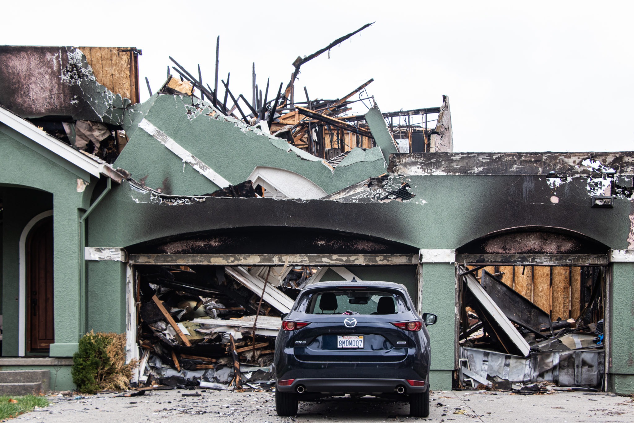 A severely fire-damaged house with a collapsed roof and charred walls is pictured, presenting a scene that might capture the interest of Berkeley Journalism students. A blue SUV is parked in front of the garage, which has significant structural damage and debris scattered around.