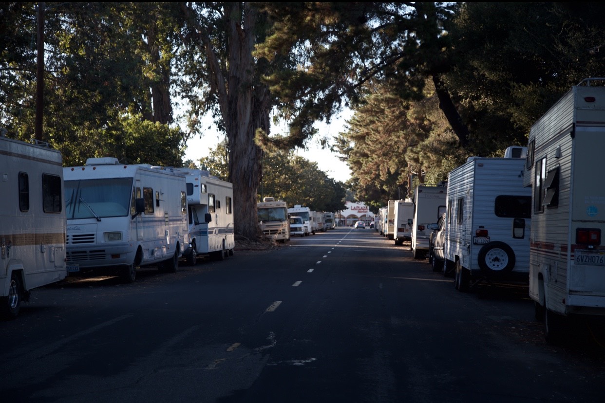 A quiet street lined with parked RVs on both sides. Tall trees provide shade, and soft sunlight filters through the leaves. The road is empty of pedestrians and other vehicles, creating a serene atmosphere reminiscent of the tranquil surroundings often captured by Berkeley Journalism students.