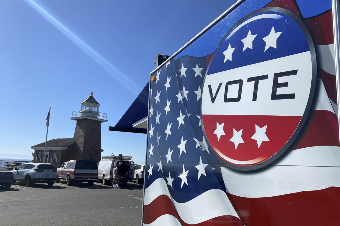 An election-themed trailer decorated with stars and stripes and a large "VOTE" sign is parked near a lighthouse on a sunny day. Several cars are parked nearby, and an American flag flutters next to the lighthouse, adding to the festive atmosphere captured by Berkeley Journalism students.