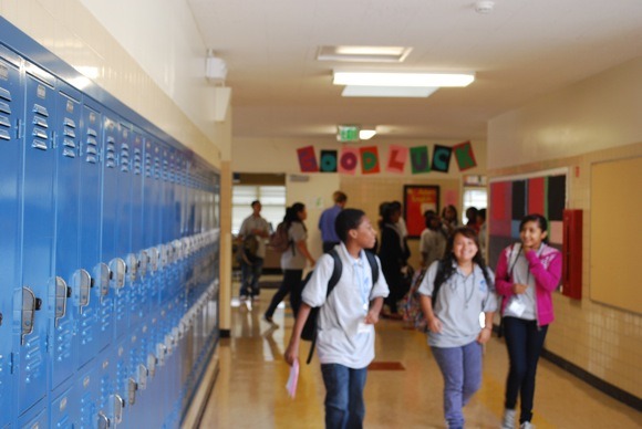 Students walk through a school hallway lined with blue lockers. Some carry backpacks and papers. A 