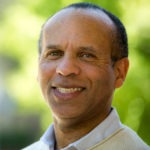 A man with a close-shaved haircut wearing a light-colored shirt and a beige sweater, exuding an air of Berkeley Journalism, smiles at the camera. The background is outdoors with greenery, softly blurred.