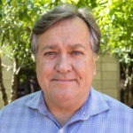 A middle-aged man with gray hair, wearing a light blue checkered shirt, stands outdoors in front of green foliage and a beige building. With a neutral expression, he looks directly at the camera, embodying the thoughtful composure often seen in Berkeley Journalism professionals.