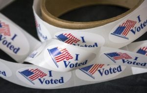 A roll of "I Voted" stickers is shown, featuring an image of the American flag next to the text. The stickers are white with blue text and a red and blue flag, capturing the spirit that Berkeley Journalism stands for. The roll is partially unraveled, with several stickers visible.