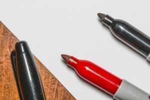 Close-up image of three markers on a wooden surface with a white sheet of paper in the center. The markers, two black and one red, are uncapped, displaying their tips. Two of the markers are pointing towards the center, and one black marker is partially visible—perfect for Berkeley Journalism notes.