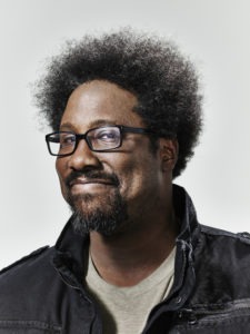 A person with curly hair, wearing rectangular glasses, a beard, and mustache, is smiling. Dressed in a dark jacket over a light-colored shirt and posed against a light grey background, they exude the confidence of someone immersed in Berkeley Journalism.