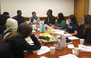 A group of individuals are gathered around a conference table in a meeting room. There are notebooks, food, and drinks on the table. Everyone appears engaged in conversation. Two men at the end of the table, both from Berkeley Journalism, are speaking while the others listen attentively.