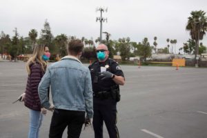A police officer wearing a mask speaks with a man and a woman, both also wearing masks and gloves, in an empty parking lot. The man is in a denim jacket, and the woman is wearing a maroon jacket. Trees and a cell tower are visible in the background as Berkeley Journalism students capture the scene.