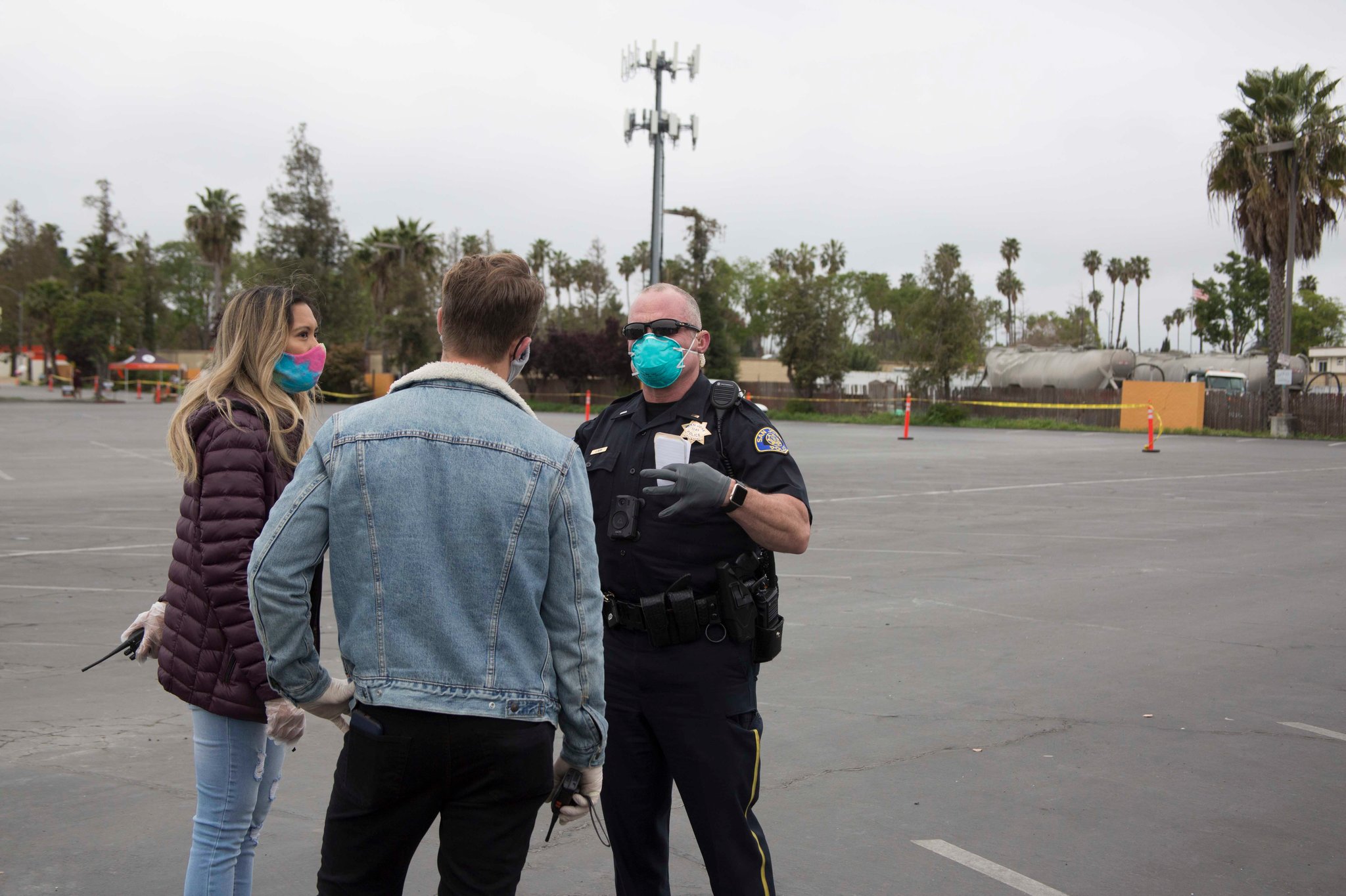 A police officer wearing a mask speaks with a man and a woman, both also wearing masks and gloves, in an empty parking lot. The man is in a denim jacket, and the woman is wearing a maroon jacket. Trees and a cell tower are visible in the background as Berkeley Journalism students capture the scene.