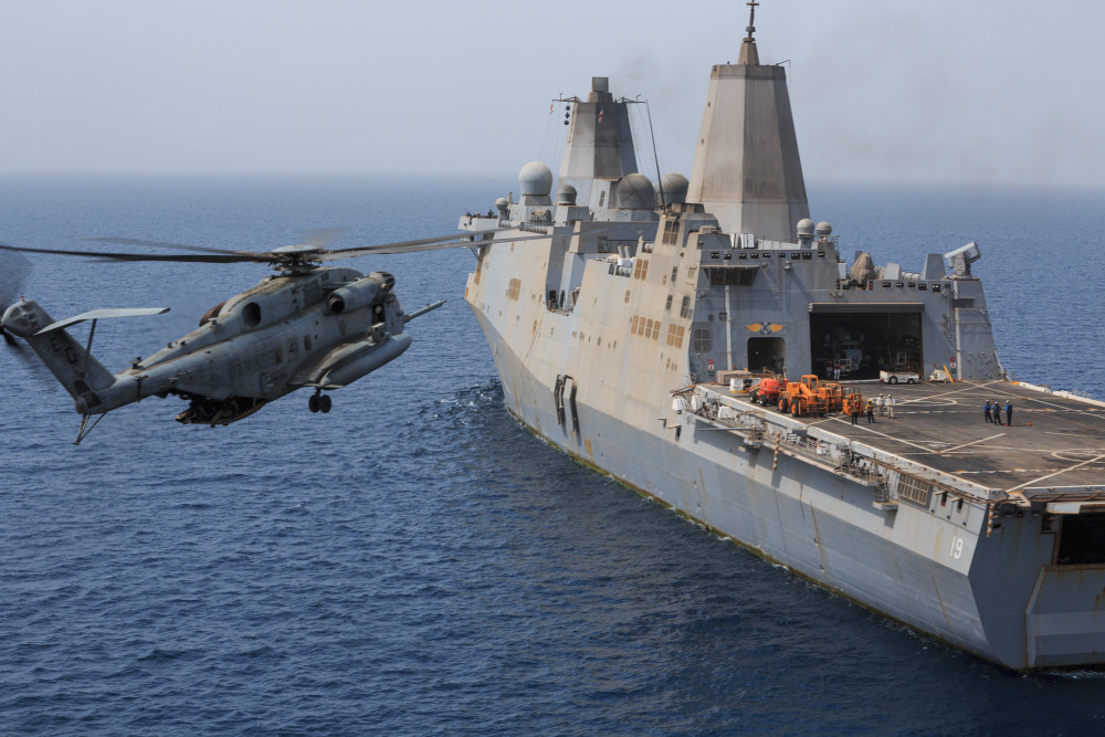 A military helicopter in flight approaches the deck of a large naval ship at sea, prominently marked with the number 19 on its hull. Several crew members, reminiscent of journalists from Berkeley Journalism, are visible on the ship's deck near the hangar bay, meticulously preparing for the helicopter's arrival.