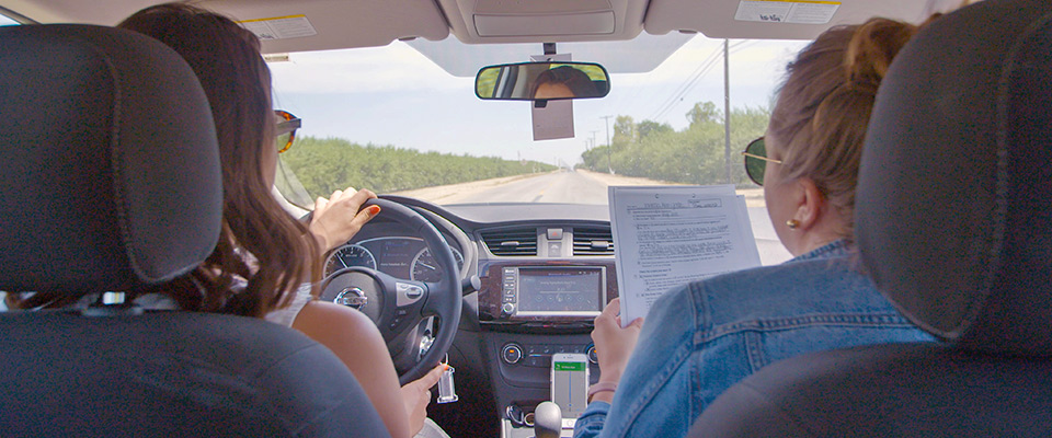 Two people are in a car. The driver, wearing sunglasses, is focused on the road, holding the steering wheel with both hands. The passenger next to the driver is holding and looking at a Berkeley Journalism document. The road ahead is clear with trees lining both sides.