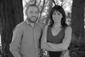 A black and white photo of a man and a woman standing outside in front of large trees. The man, possibly a Berkeley Journalism alum, is smiling and wearing a checkered shirt with rolled-up sleeves. The woman is also smiling with her arms crossed and is wearing a patterned dress.