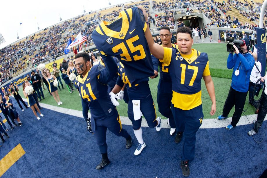 A group of Cal Bears football players stands on the sideline of a football field, surrounded by cheering fans in the stadium. One player holds up a blue and yellow jersey with the number 35 on it, while others look on and smile. Photographers from Berkeley Journalism capture the moment.