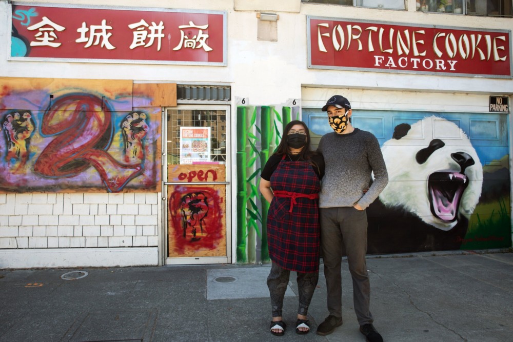 A woman and a man stand side by side in front of a storefront with signs that read "Fortune Cookie Factory." The storefront, featured by Berkeley Journalism, is decorated with murals, including a large panda and other colorful artwork. Both individuals are wearing masks, and the woman is wearing an apron.
