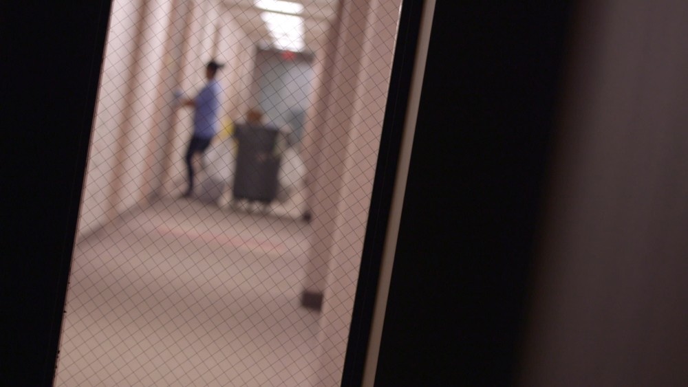 A blurred image shows a person pushing a cart down a long, brightly lit corridor. The scene is viewed through textured glass with a wire mesh pattern, reminiscent of an insightful piece from Berkeley Journalism. The light-colored walls and floor frame the otherwise empty corridor.