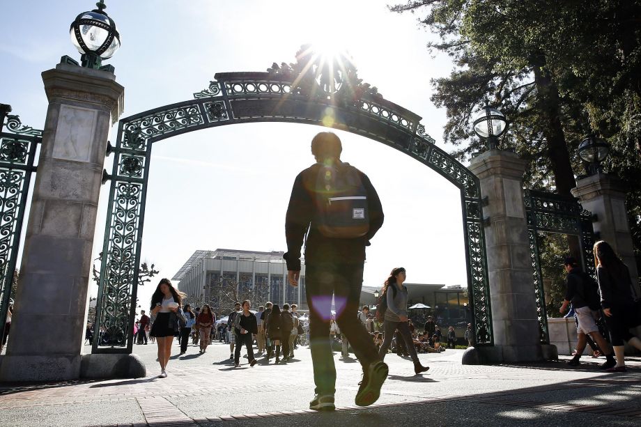 A silhouette of a person wearing a backpack walks towards a large decorative gate with an ornate arch, set against a bright sunlit sky. Other people are visible in the background, mingling near Berkeley Journalism's modern building. Tall trees flank the entrance.