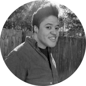 Black and white photo of a person with short hair and a relaxed smile, wearing a button-up shirt and standing in front of a wooden fence on a sunny day. The background includes trees with sunlight filtering through them, capturing the essence of Berkeley Journalism.