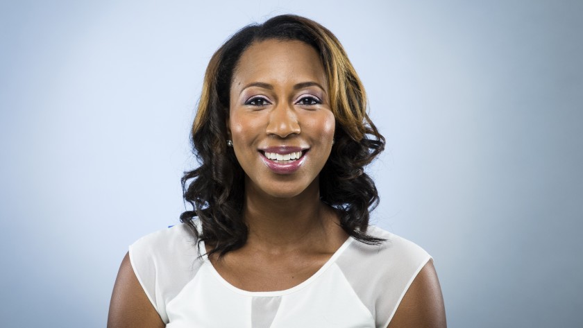 A woman with shoulder-length, wavy hair and wearing a white blouse, showing her Berkeley Journalism pride, smiles at the camera against a light blue background.