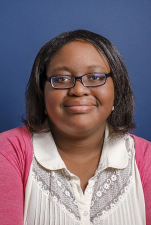 A person with shoulder-length hair and glasses smiles slightly at the camera. They are wearing a white lace-embellished blouse under a pink cardigan, embodying the vibrant energy of Berkeley Journalism. The background is a solid blue color.