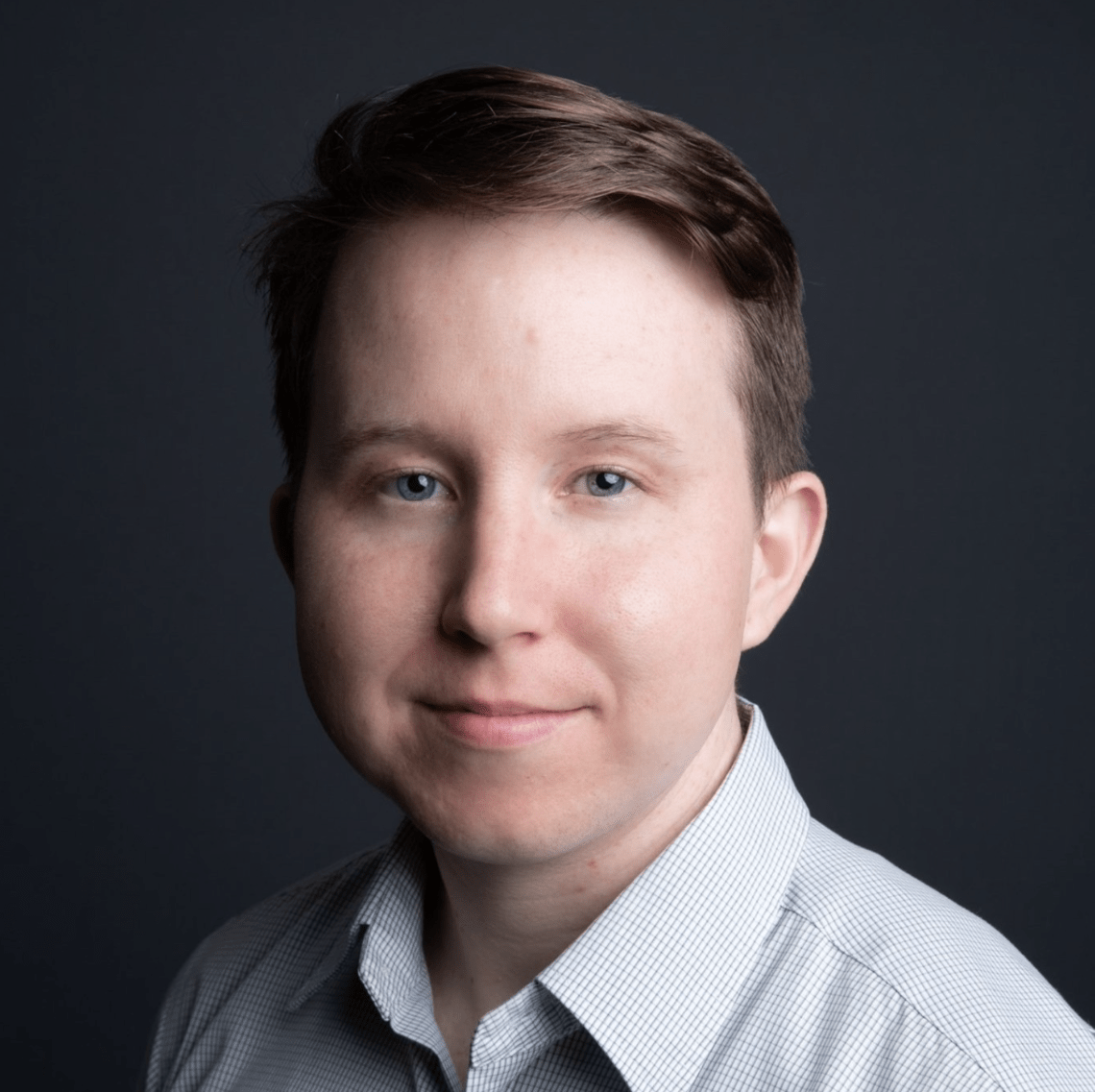 A person with short brown hair is looking directly at the camera with a neutral expression. They are wearing a light gray, checkered collared shirt, reminiscent of a thoughtful Berkeley Journalism student. The background is dark, enhancing the contrast with their light-colored attire.